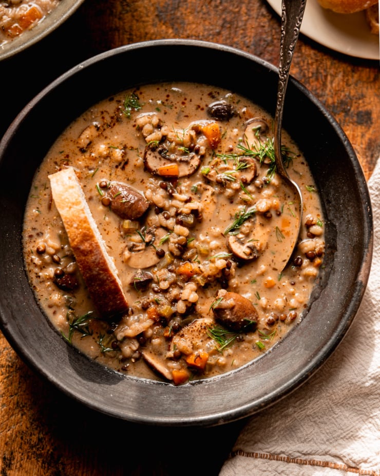 An overhead shot shows a bowl of vegan mushroom barley soup. The soup also features black lentils and fresh dill. A spoon is sticking out of the bowl of soup and a linen napkin is nestled beside the bowl. A slice of bread is perched right in the bowl of soup as well.