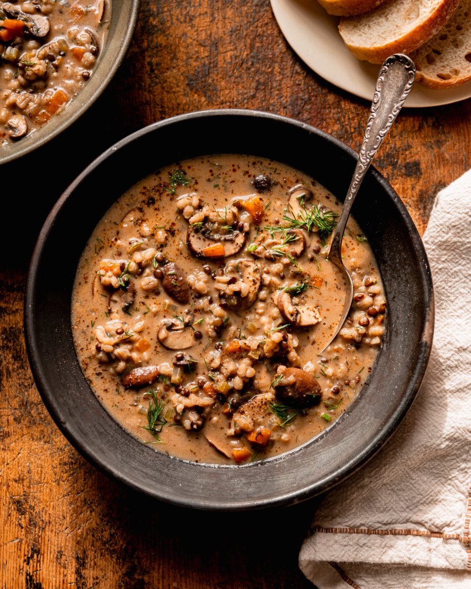 An overhead shot shows a bowl of vegan mushroom barley soup. The soup also features black lentils and fresh dill. A spoon is sticking out of the bowl of soup and a linen napkin is nestled beside the bowl.