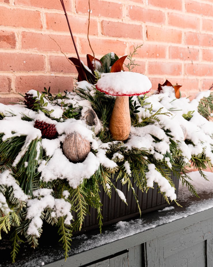 A head-on, angled shot shows a holiday arrangement of winter greens, ornaments and branches. The arrangement is covered in a fluffy layer of snow.