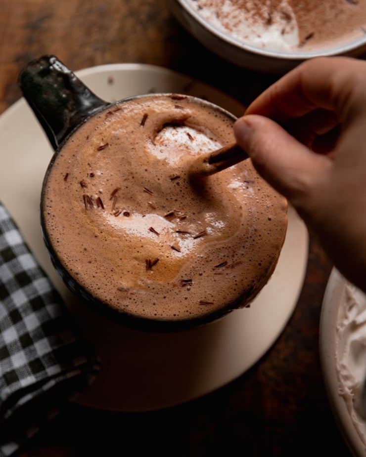 An overhead shot shows a hand using a cinnamon stick to stir up a mug of vegan orange mocha.