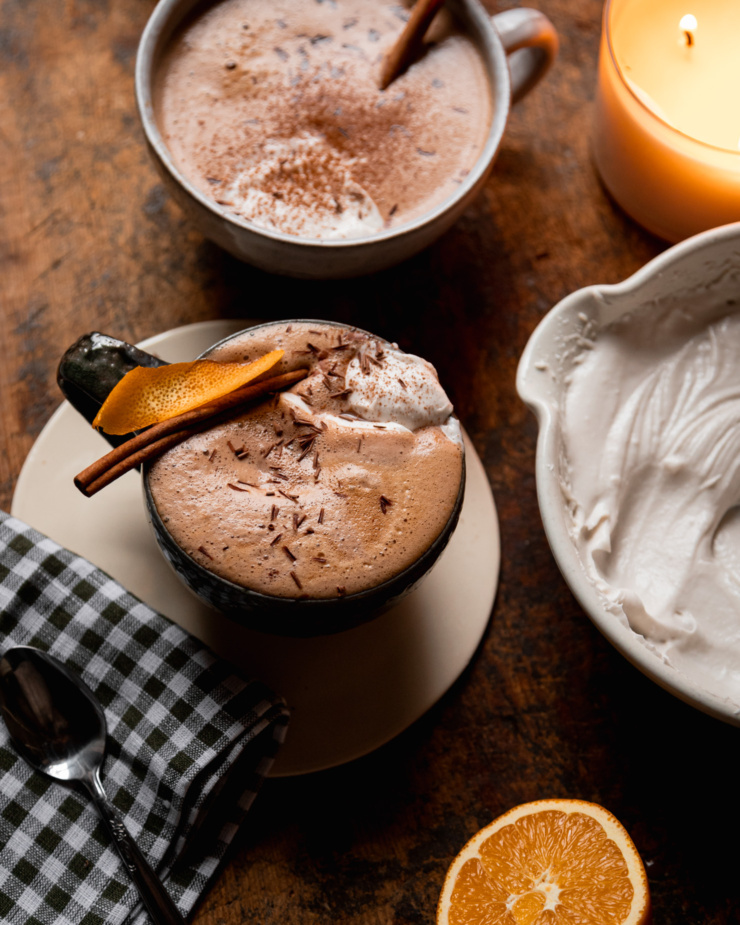 A overhead shot shows an orange mocha in a mug. The mocha is garnished with orange peel and a cinnamon stick and topped with coconut whipped cream and chocolate shavings. A halved orange and bowl of coconut whipped cream is nearby.