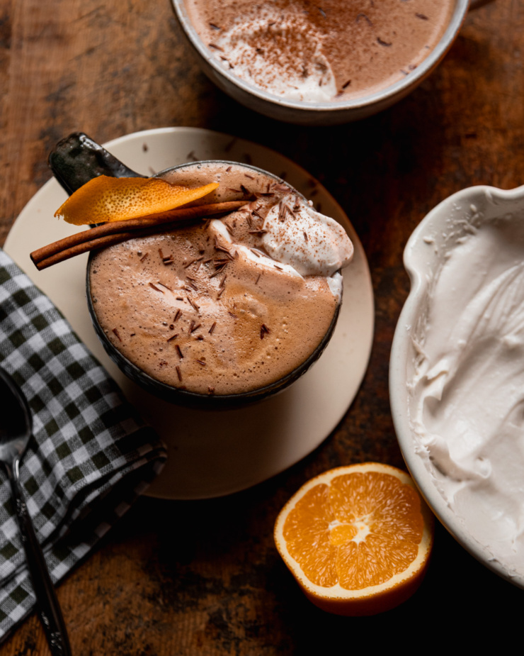 A overhead shot shows an orange mocha in a mug. The mocha is garnished with orange peel and a cinnamon stick and topped with coconut whipped cream and chocolate shavings. A halved orange and bowl of coconut whipped cream is nearby.