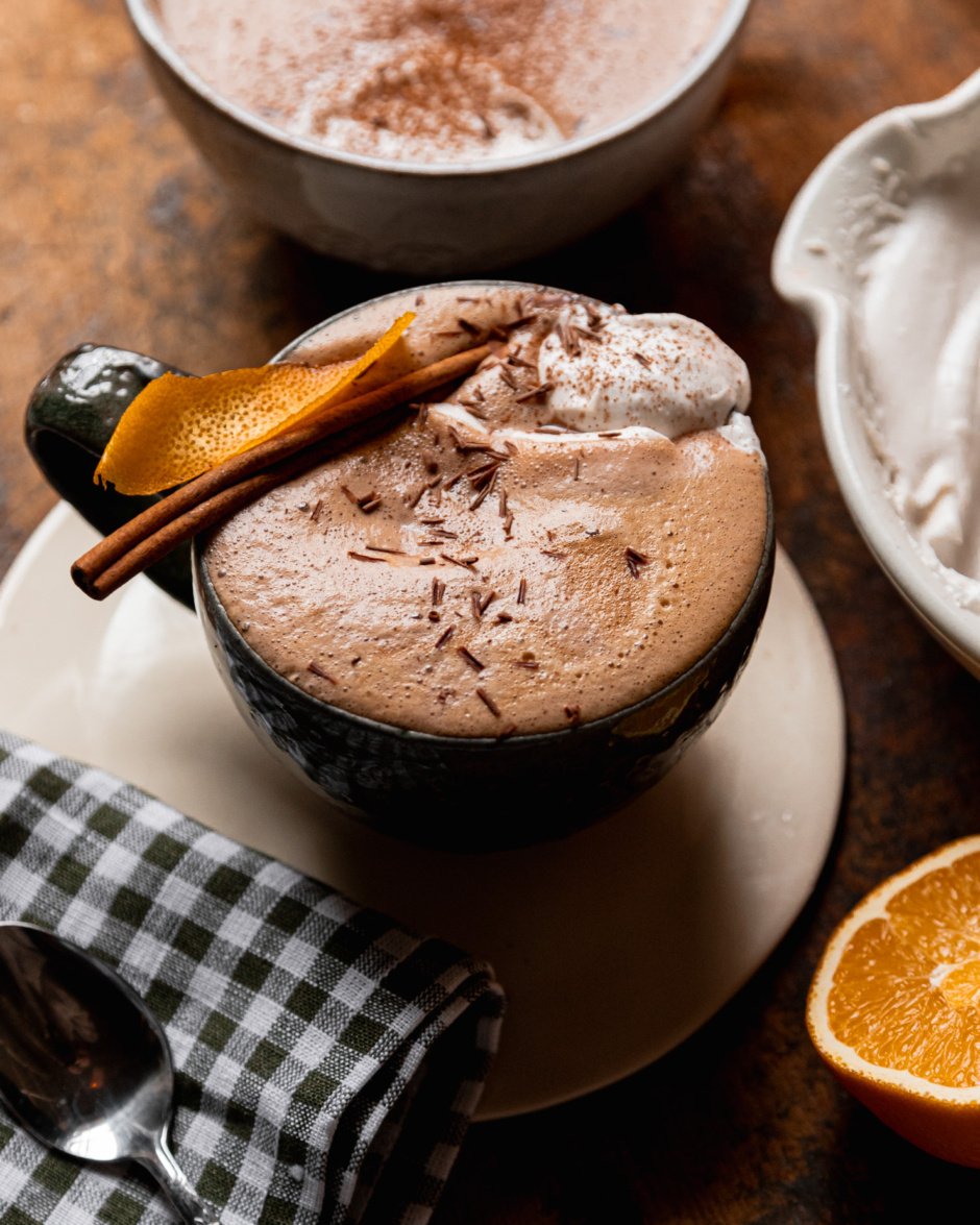 A 3/4 angle shot shows an orange mocha in a mug. The mocha is garnished with orange peel and a cinnamon stick and topped with coconut whipped cream and chocolate shavings.