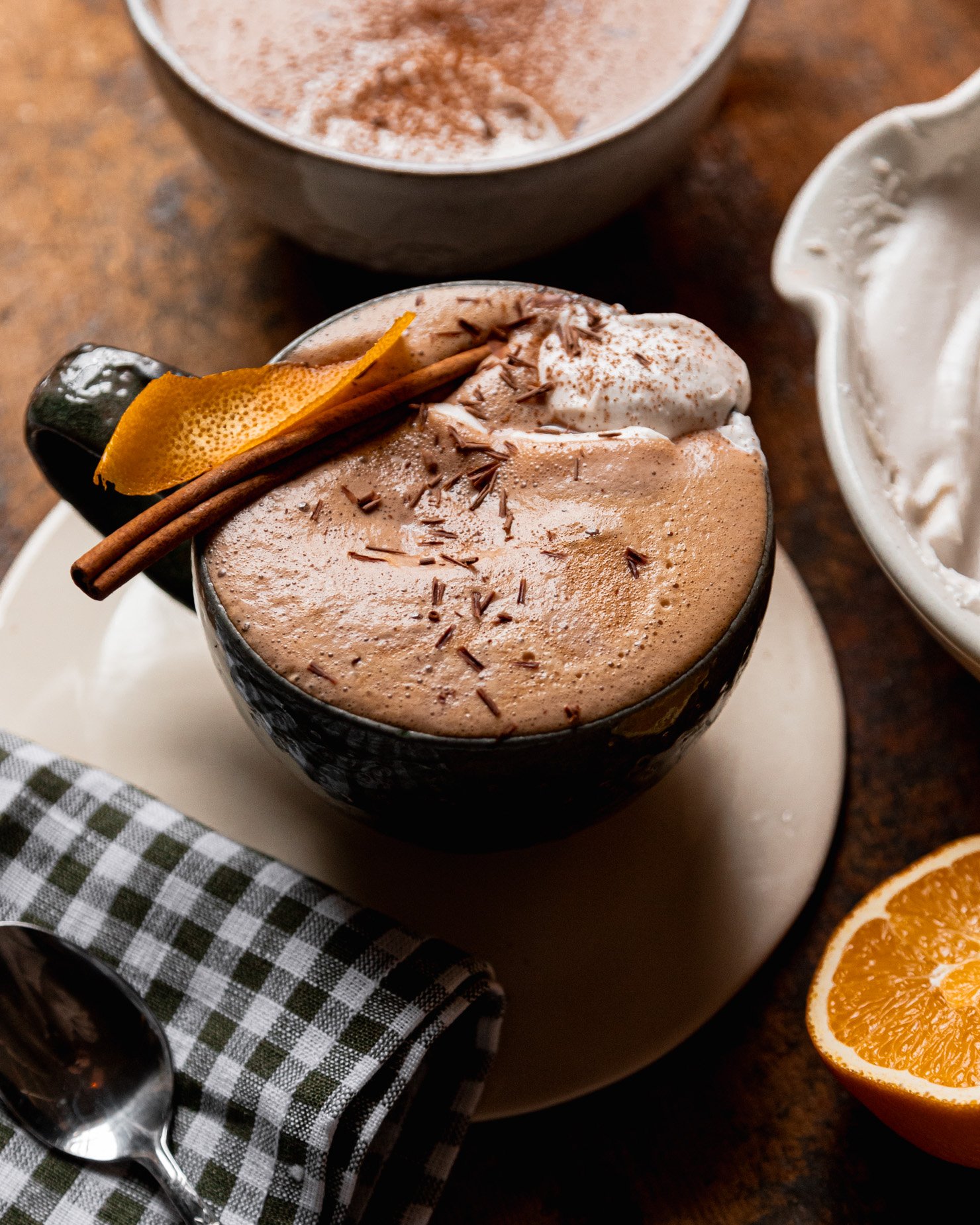 A 3/4 angle shot shows an orange mocha in a mug. The mocha is garnished with orange peel and a cinnamon stick and topped with coconut whipped cream and chocolate shavings.