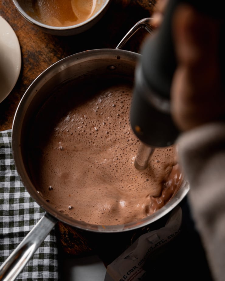 An overhead shot shows a hand using an immersion blender to froth up an orange mocha milk mixture in a stainless steel saucepan.