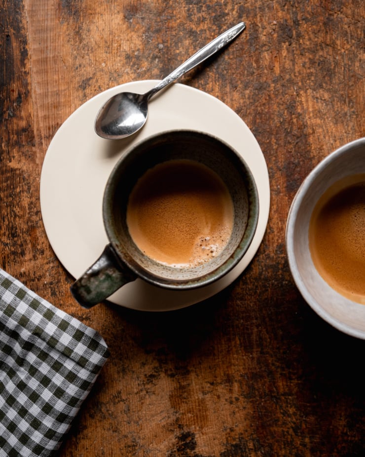 An overhead shot shows two cups with two shots of espresso in them, against a rough wood background.