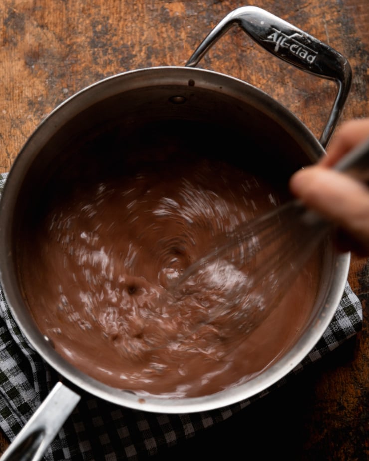 An overhead shot shows a hand using a whisk to bring a chocolate milk mixture in a saucepan. There are blurs as the whisking is quite rapid.