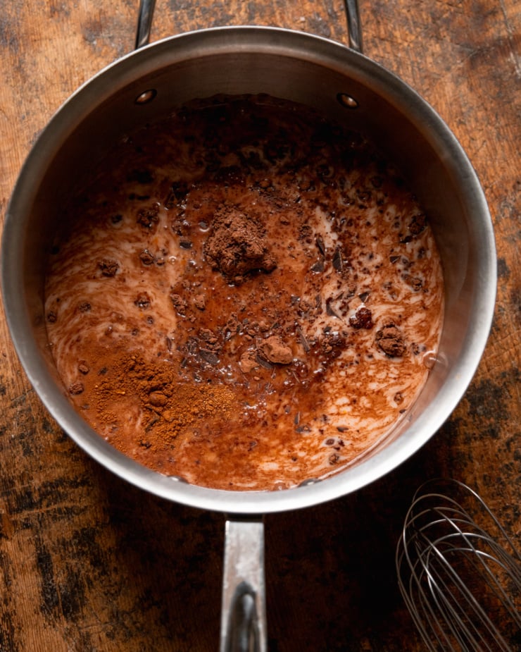 An overhead shot shows a stainless saucepan filled with non-dairy milk, cocoa powder, spices, chopped chocolate, and more.