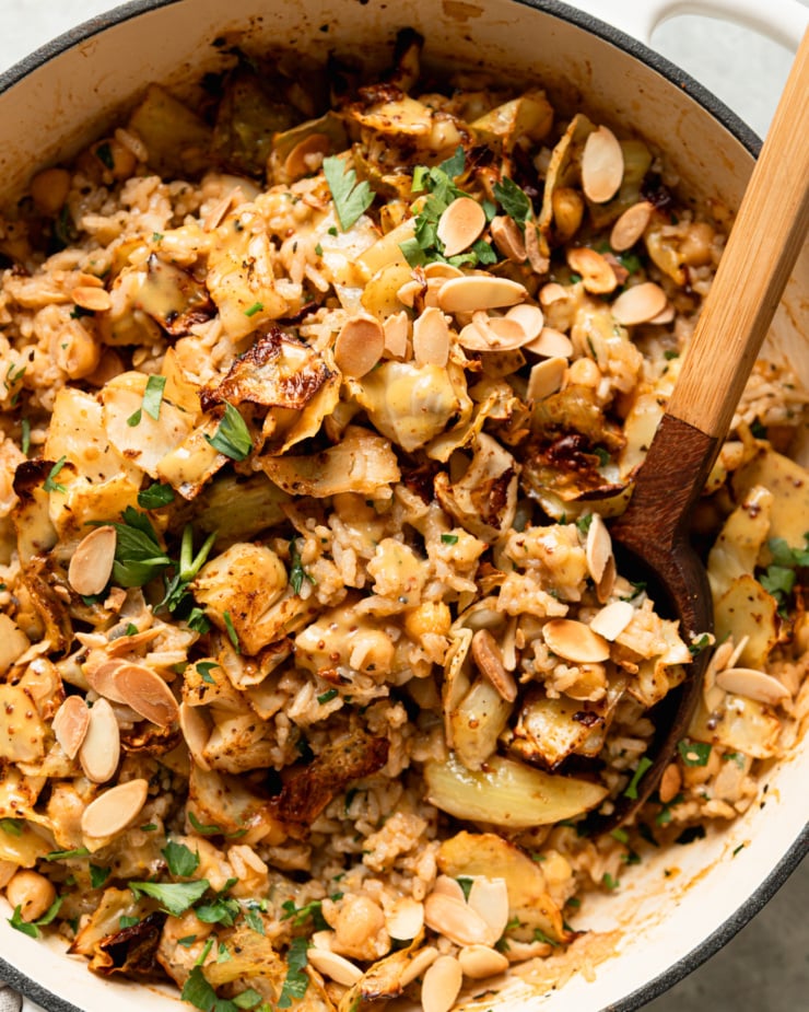 An up close, overhead shot shows a braiser pot filled with a mixture of chickpeas and rice, crispy roasted cabbage, chopped parsley, drizzles of sweet mustard dressing, and toasted sliced almonds. A spoon is sticking out of the pot.