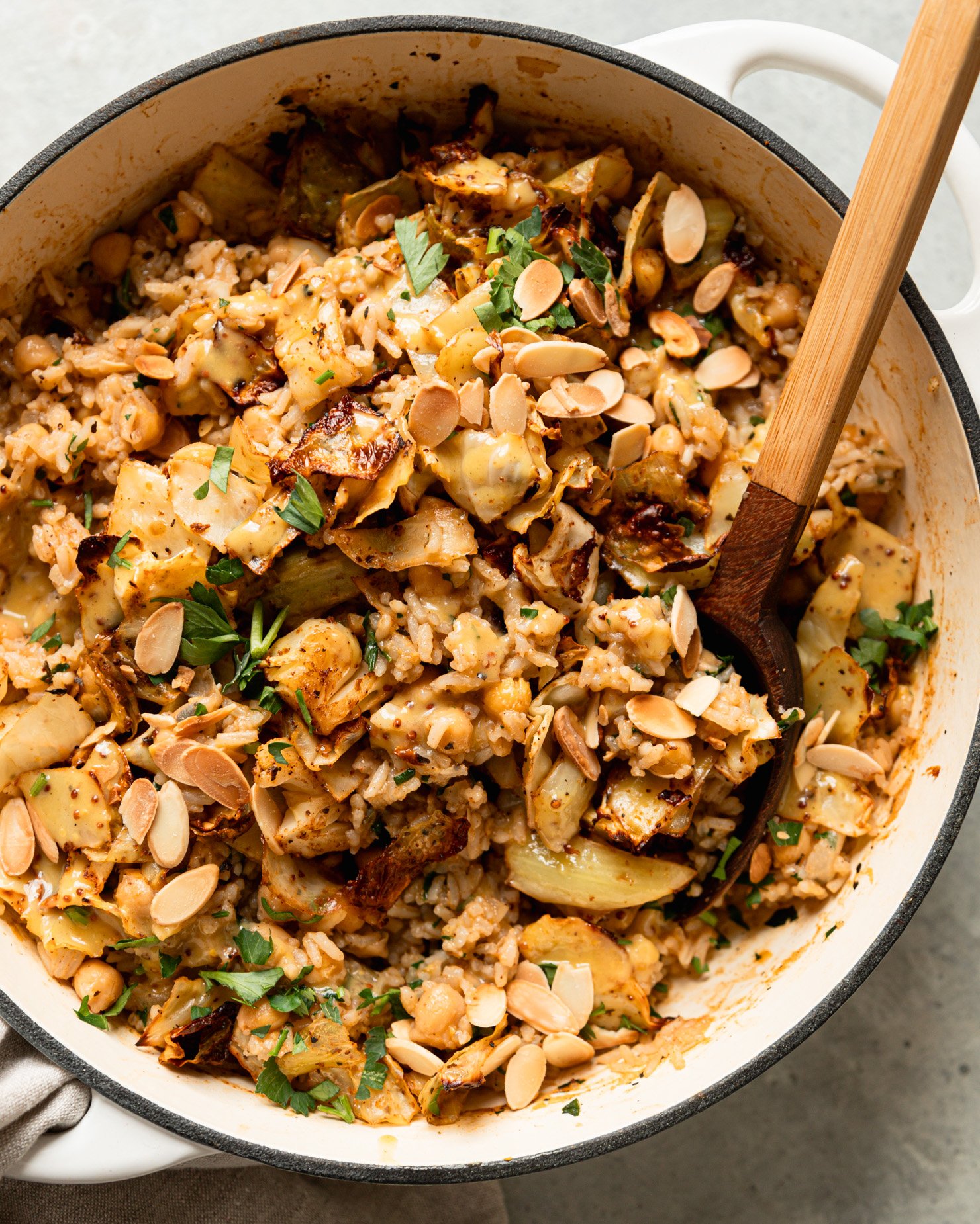 An overhead shot shows a braiser pot filled with a mixture of chickpeas and rice, crispy roasted cabbage, chopped parsley, drizzles of sweet mustard dressing, and toasted sliced almonds. A spoon is sticking out of the pot.