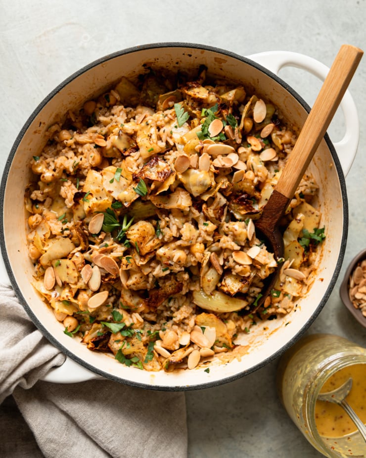 An overhead shot shows a braiser pot filled with a mixture of chickpeas and rice, crispy roasted cabbage, chopped parsley, drizzles of sweet mustard dressing, and toasted sliced almonds. A spoon is sticking out of the pot and a jar of the dressing is nearby.