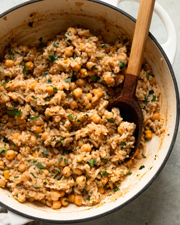 An overhead shot shows a braiser pot filled with a spiced rice and chickpea mixture. Chopped parsley has been stirred throughout.