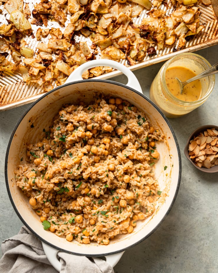 An overhead shot shows a pot of chickpeas and rice, a tray of roasted crispy cabbage, a jar of sweet mustard dressing, and a small bowl of toasted sliced almonds.
