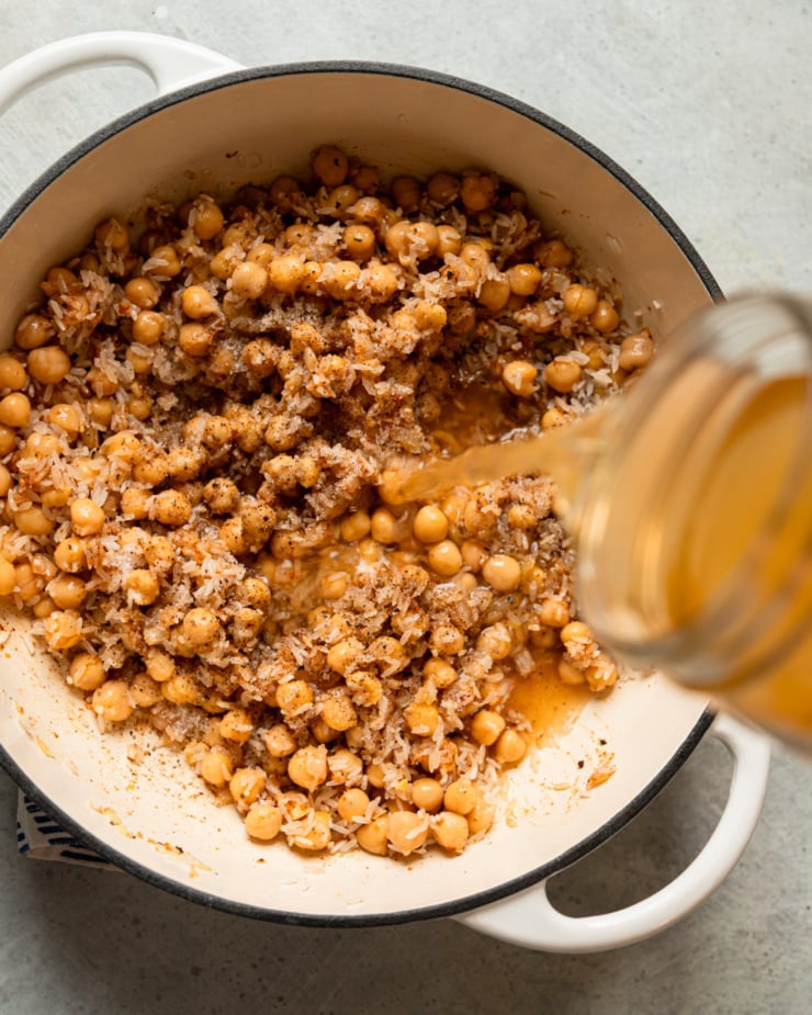 An overhead shot shows vegetable stock being poured into a pot of chickpeas, onions, and uncooked rice.