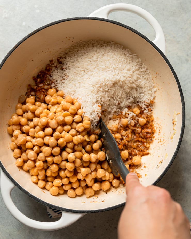 An overhead shot shows a spiced sautéed onion mixture in a pot with raw rice and canned chickpeas on top. A hand is using a spatula to stir it all together.