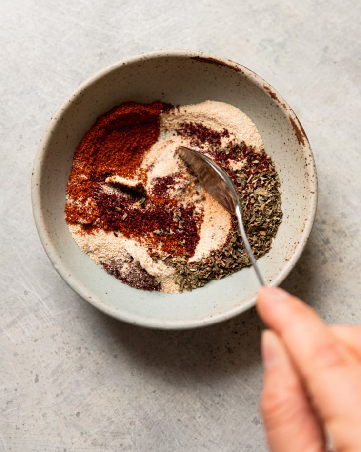 An overhead shot shows a hand using a spoon to stire some spices together in a bowl.