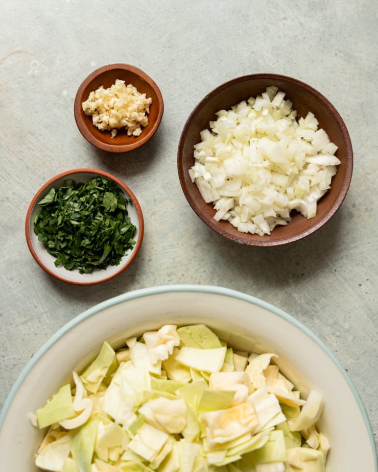 An overhead shot shows bowls containing: minced garlic, chopped onion, chopped cabbage, and chopped parsley.