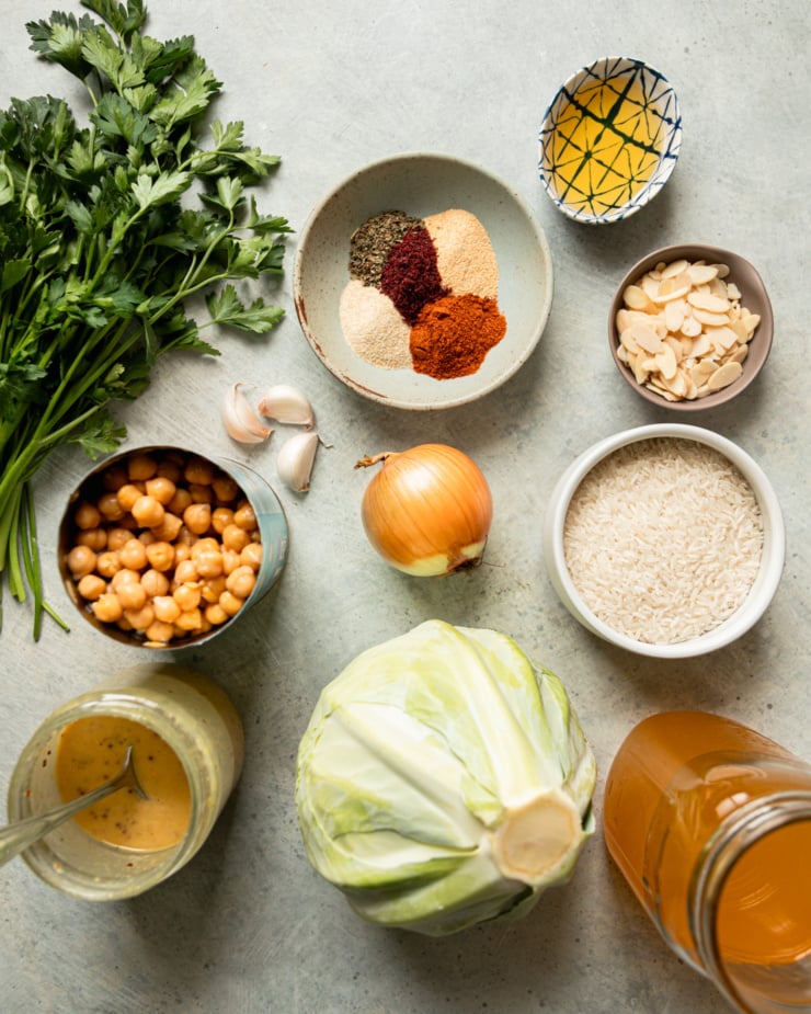 An overhead shot shows ingredients for a vegan dinner recipe: parsley, spices, olive oil, sliced almonds, onion, jasmine rice, vegetable stock, cabbage, sweet mustard dressing, canned chickpeas, and garlic cloves.