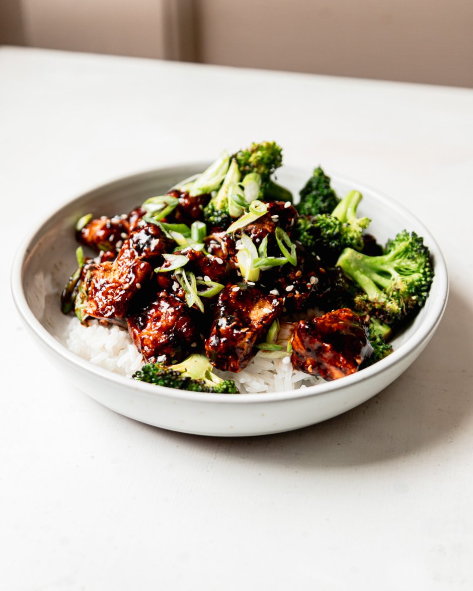A head-on shot shows a bowl of rice topped with crunchy and glossy nuggets of teriyaki tofu and cooked broccoli. The dish is garnished with sliced green onions and sesame seeds.