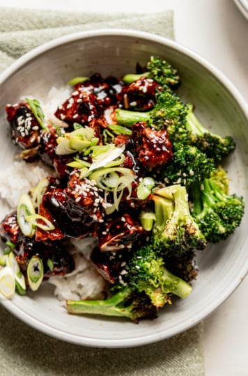 An overhead shot shows a bowl of rice topped with crunchy and glossy nuggets of teriyaki tofu and cooked broccoli. The dish is garnished with sliced green onions and sesame seeds.