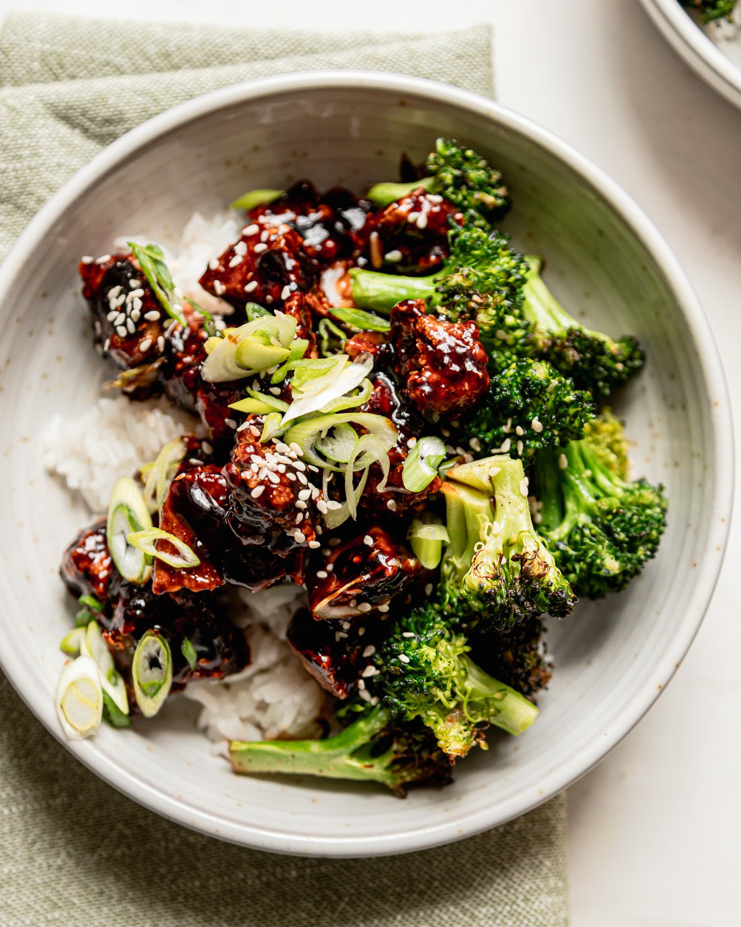 An overhead shot shows a bowl of rice topped with crunchy and glossy nuggets of teriyaki tofu and cooked broccoli. The dish is garnished with sliced green onions and sesame seeds.