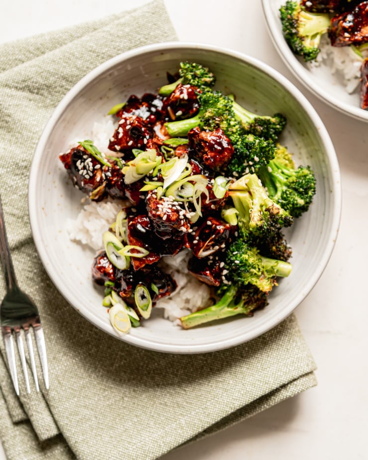 An overhead shot shows a bowl of rice topped with crunchy and glossy nuggets of teriyaki tofu and cooked broccoli. The dish is garnished with sliced green onions and sesame seeds. A napkin and fork are seen nearby.