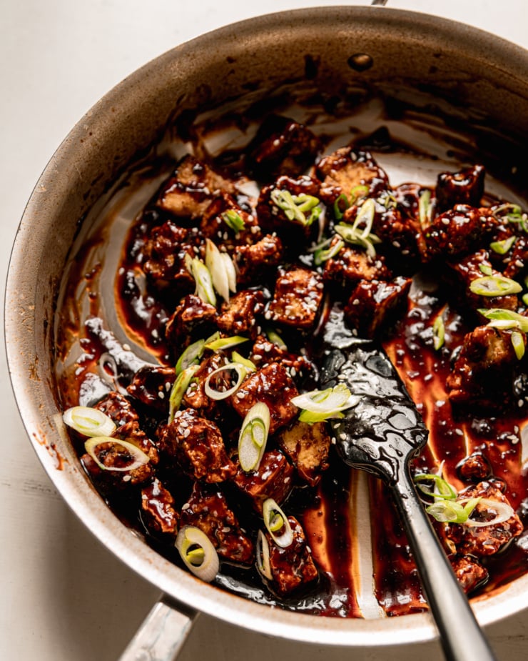 A 3/4 angle shot shows teriyaki sauce-coated tofu pieces in a skillet, garnished with sliced green onions and sesame seeds. A spatula sticks out of the skillet.