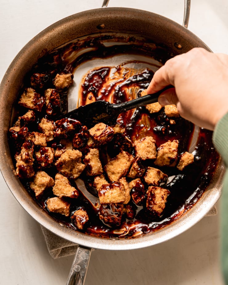 An overhead shot shows a hand using a spatula to stir some crunchy air fried tofu pieces into glossy and thick homemade teriyaki sauce in a skillet.