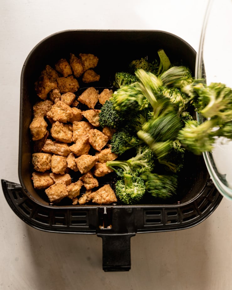 An overhead shot shows crunchy tofu bits in an air fryer basket. Broccoli florets are being poured into the basket as well.