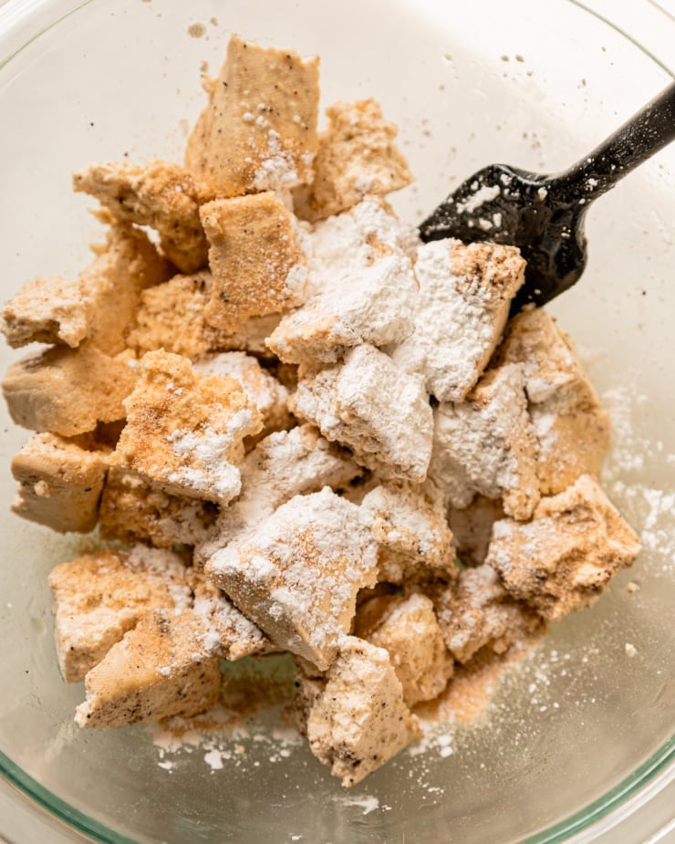 An overhead shot shows torn firm tofu pieces in a bowl that have been dusted with spices, pepper, and cornstarch. A spatula is sticking out of the bowl.