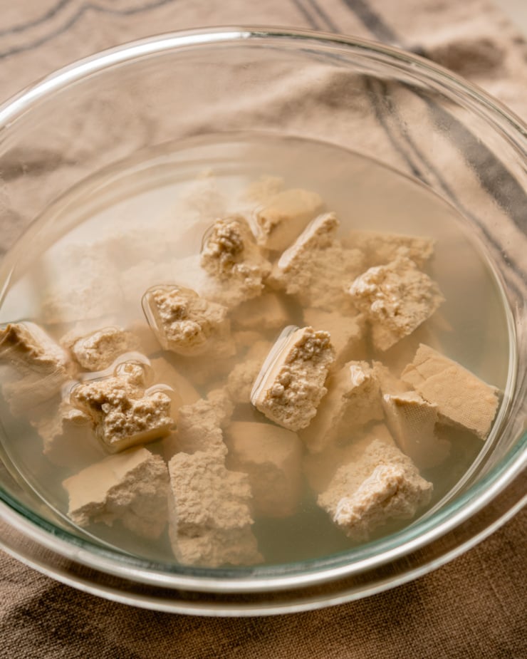 A 3/4 angle image shows torn firm tofu pieces submerged in a bowl of hot salted water.
