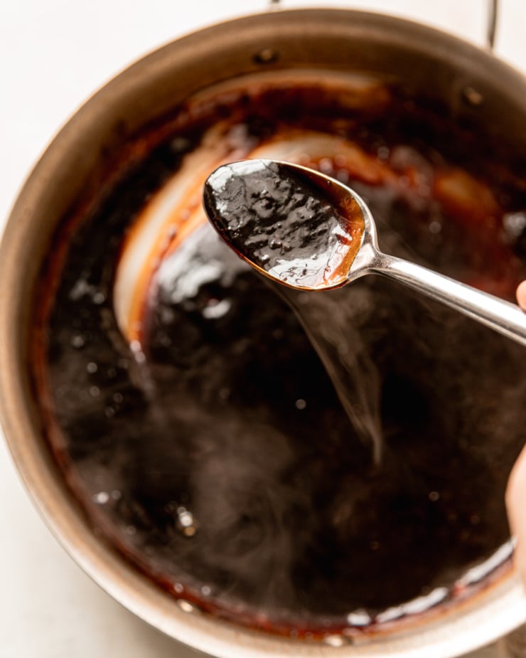 An overhead shot shows a spoon filled with glossy homemade teriyaki sauce. The spoon is held over a skillet of the steaming sauce.