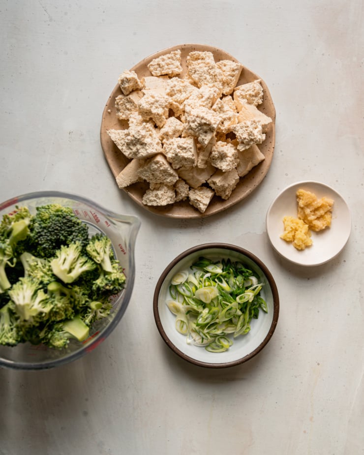 An overhead shot shows a plate of torn firm tofu pieces, minced ginger and garlic, sliced green onions, and broccoli florets.
