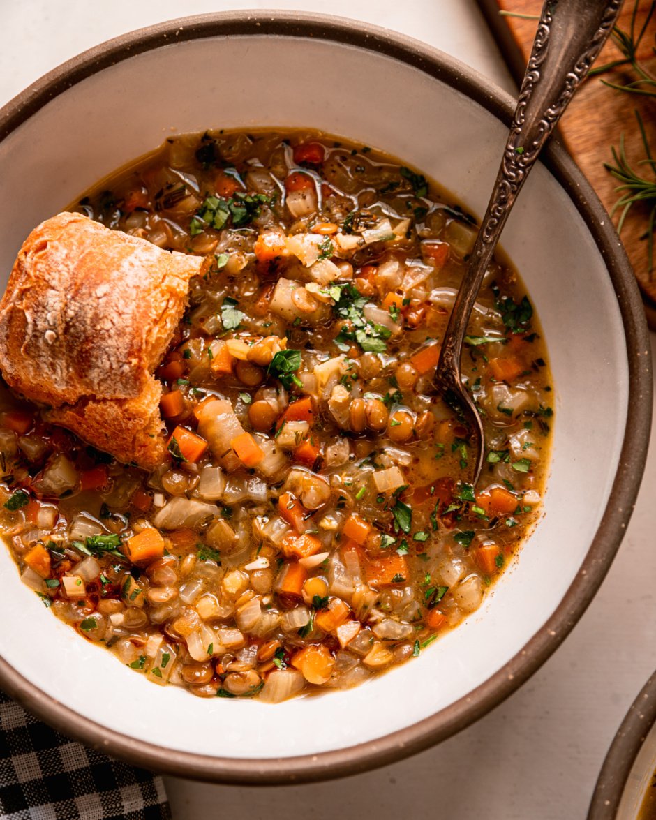 An overhead shot shows a bowl of lentil vegetable soup with a crust of bread perched on top. The soup is finished with finely chopped parsley. A spoon is sticking out of the bowl.
