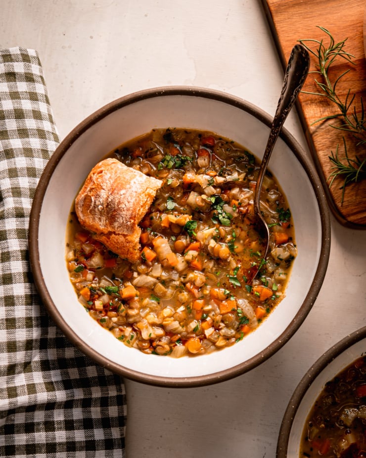 An overhead shot shows a bowl of lentil vegetable soup with a crust of bread perched on top. The soup is finished with finely chopped parsley. A spoon is sticking out of the bowl.