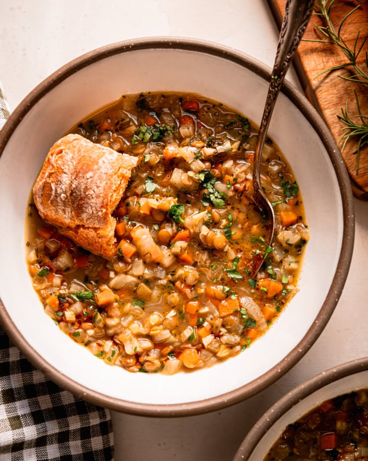 An overhead shot shows a bowl of lentil vegetable soup with a crust of bread perched on top. The soup is finished with finely chopped parsley. A spoon is sticking out of the bowl.