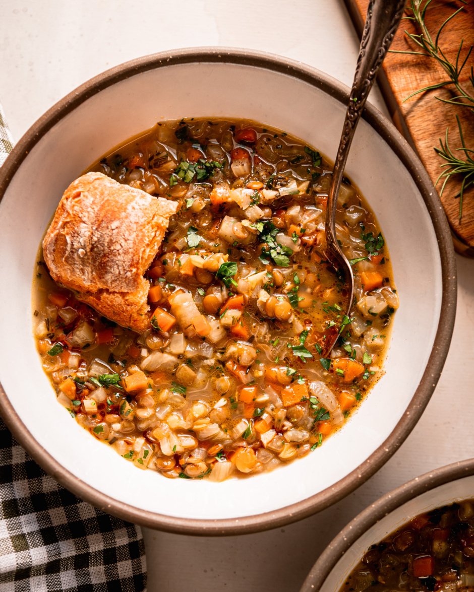 An overhead shot shows a bowl of lentil vegetable soup with a crust of bread perched on top. The soup is finished with finely chopped parsley. A spoon is sticking out of the bowl.