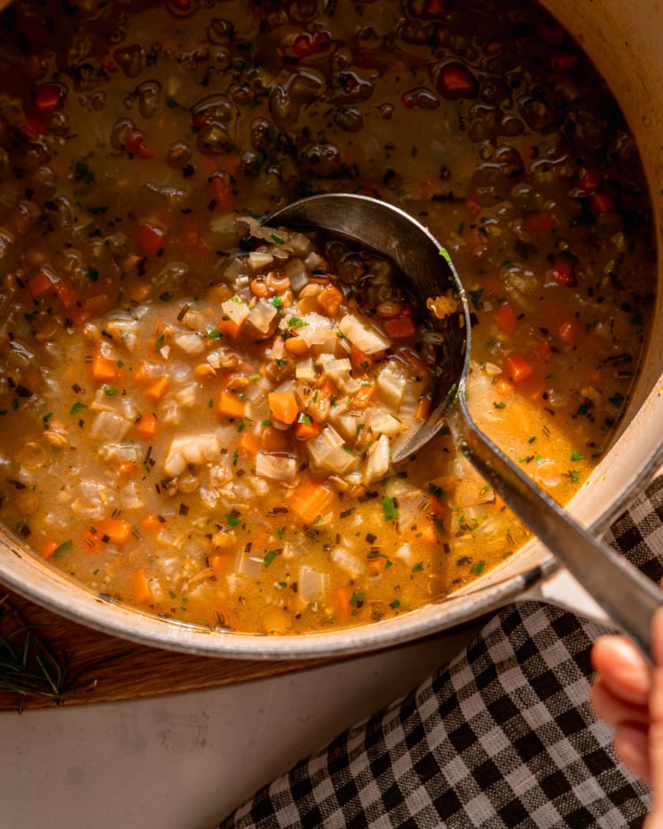 An overhead shot shows ladle emerging from a pot of lentil vegetable soup. The ladle portion is in harsh, direct sunlight.