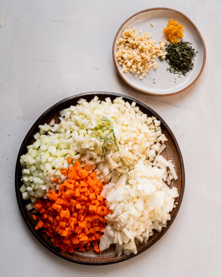 An overhead shot shows two plates. One has: minced garlic, lemon zest, and chopped rosemary. The other has: chopped fennel, carrots, celery, and onions.