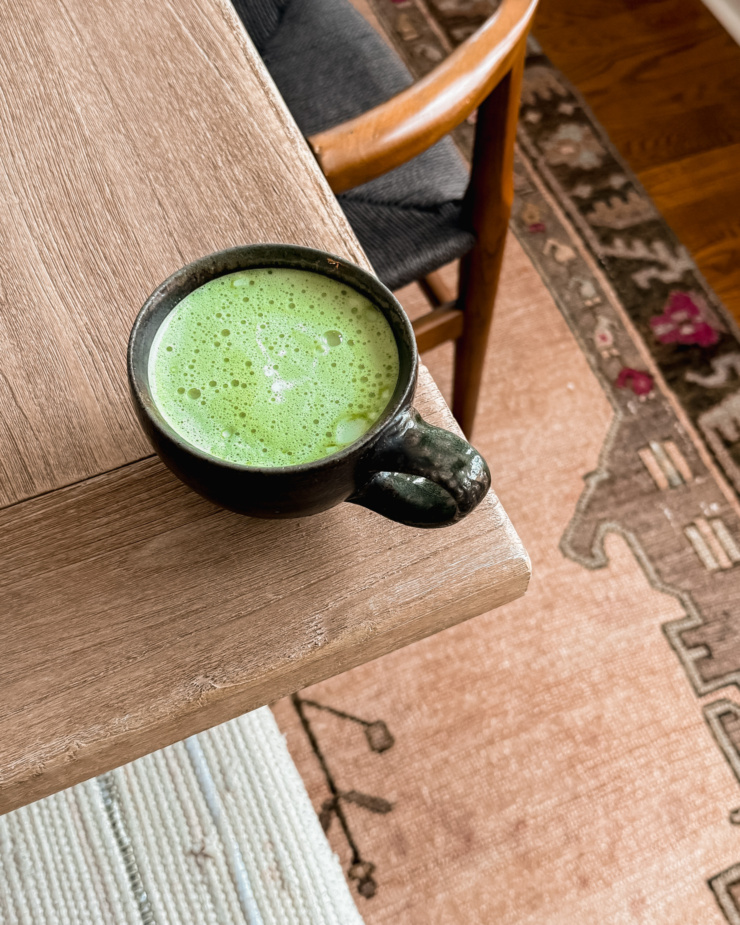 An overhead shot shows a matcha latte in a wide cup on the corner of a table surface. An ornate carpet is seen on the floor below.