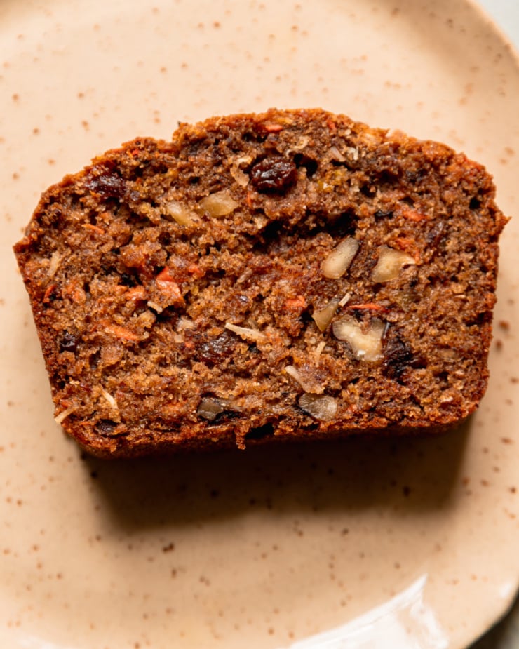 An up close, overhead shot shows a slice of morning glory bread, studded with raisins, shredded carrot and apple, coconut, and chopped walnuts.