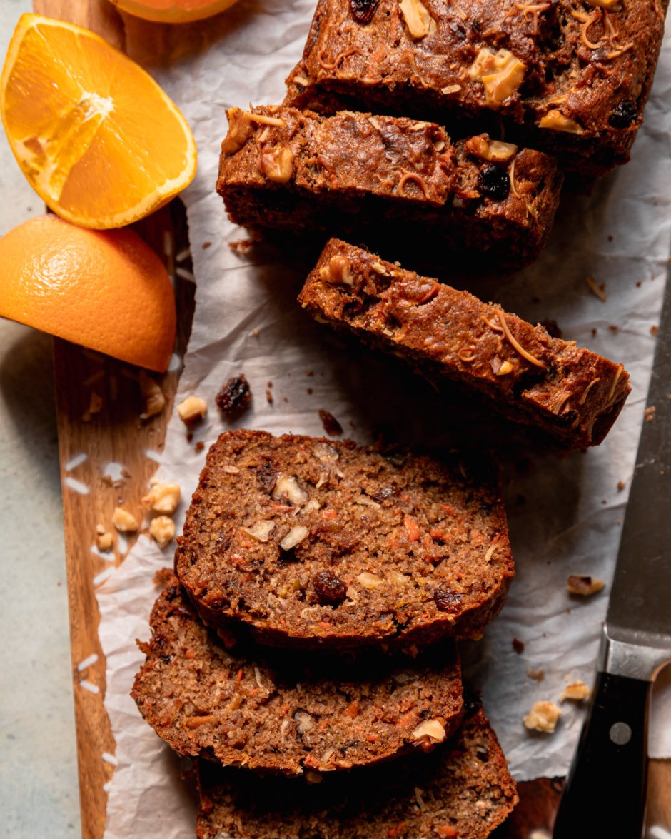 An overhead shot shows a sliced morning glory bread on a parchment-lined cutting board. Orange slices are seen nearby.