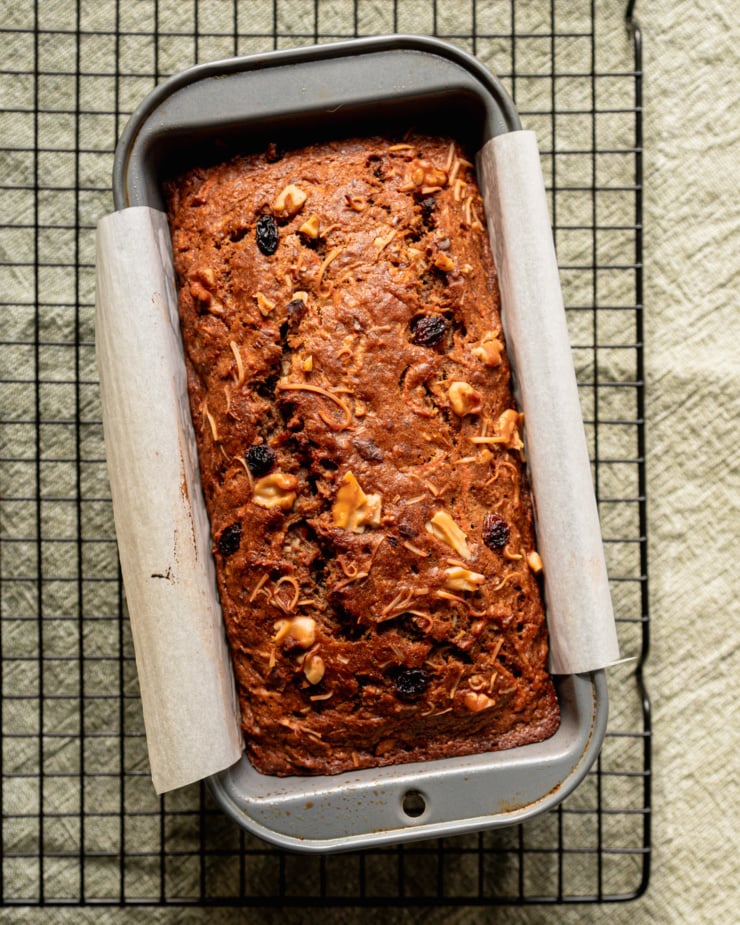 An overhead shot shows a freshly baked morning glory bread in a a parchment-lined loaf pan. The loaf is on a cooling rack.