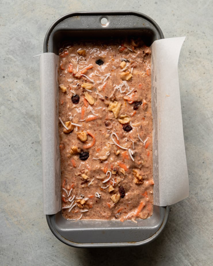 An overhead shot shows morning glory loaf batter in a parchment-lined loaf pan. The raw batter is topped with a bit of chopped walnuts, raisins, and shredded coconut for garnish.