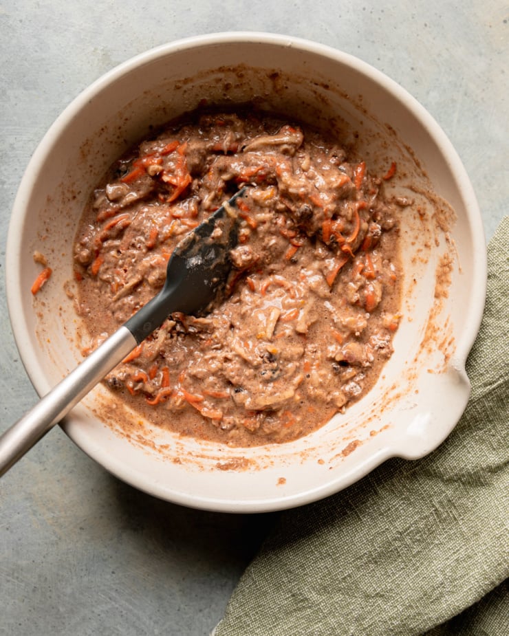 An overhead shot shows a loaf cake batter with shredded carrots and apples in a mixing bowl with a spatula sticking out.