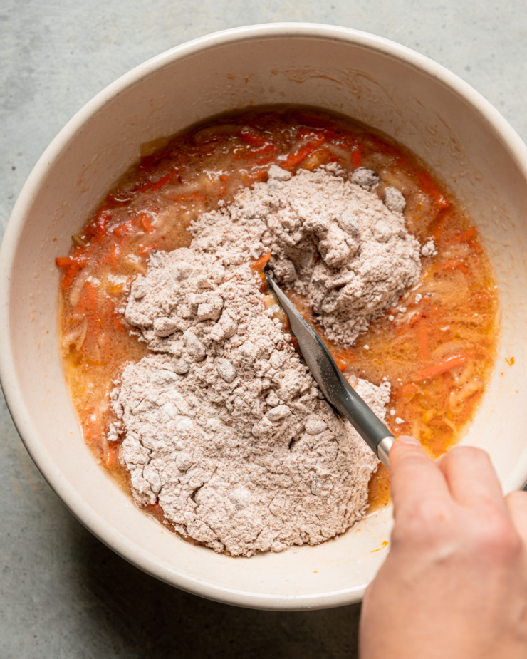 An overhead shot shows a whole wheat flour mixture added to a liquid mixture with carrots in a mixing bowl. A hand is holding a spatula in the bowl, ready to stir everything together.