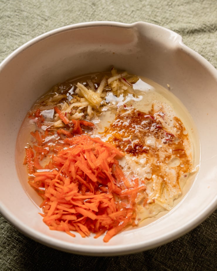 An overhead shot shows a mixing bowl filled with grated carrots and apple, soy milk, oil, maple syrup, vanilla extract, and orange juice.
