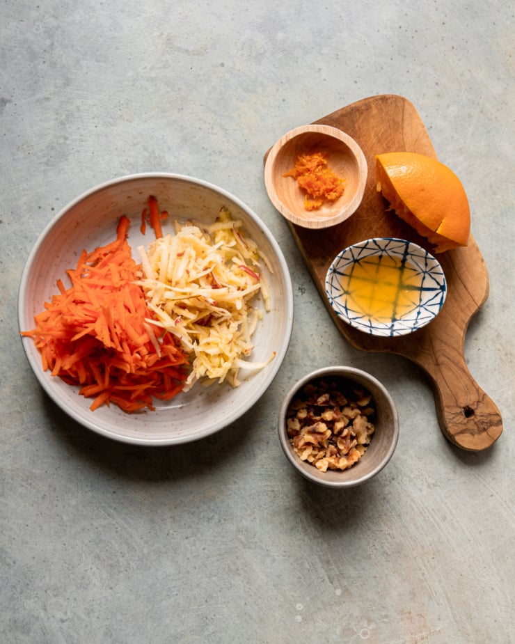 An overhead shot shows a bowl with grated carrots and apple, a small bowl with orange zest, a small bowl with orange juice, a squeezes orange half, and some chopped walnuts in a bowl.