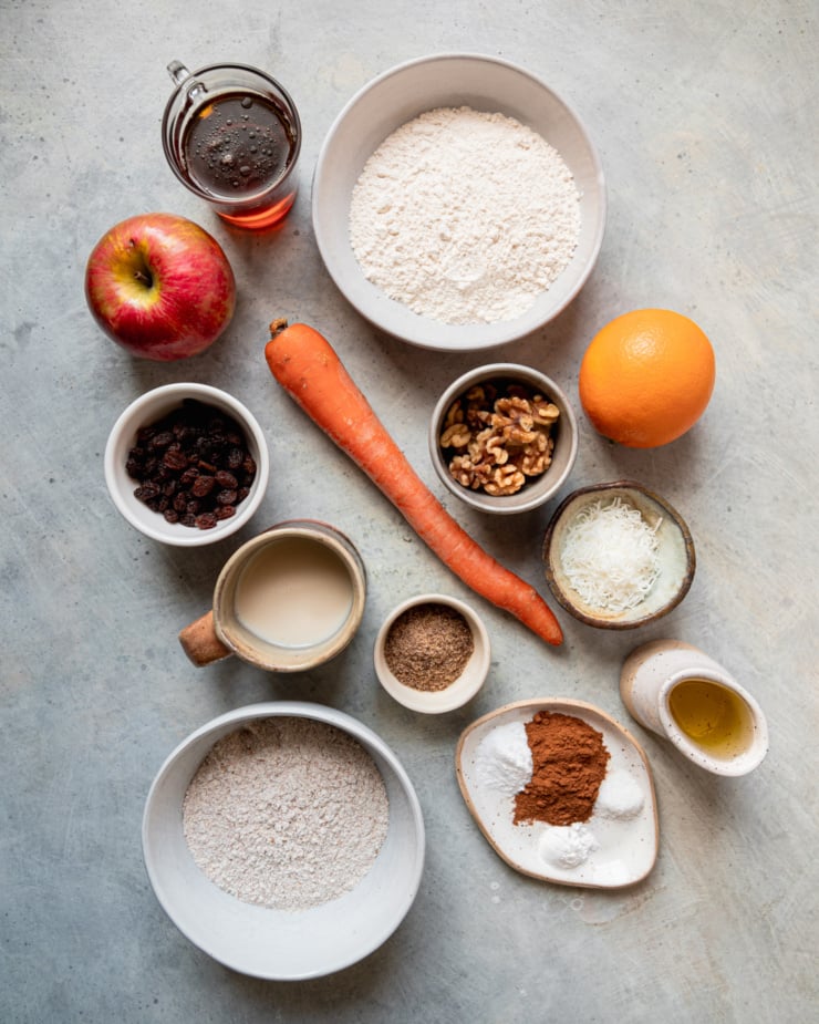 An overhead shot shows ingredients needed for a morning glory bread recipe: all-purpose flour, orange, walnuts, coconut, sunflower oil, cinnamon, salt, baking powder, baking soda, whole wheat flour, ground flax, soy milk, raisins, carrot, apple, and maple syrup.