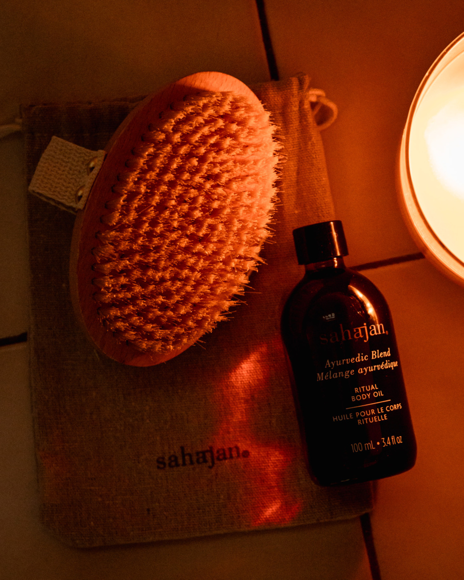 An overhead shot shows a dry brush and bottle of body oil in candlelight on top of tiles.